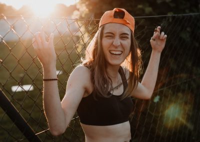 Glückliches, lachendes Skate Girl mit Kappe bei Sonnenuntergang mit einem Aachener Zaun im Hintergrund